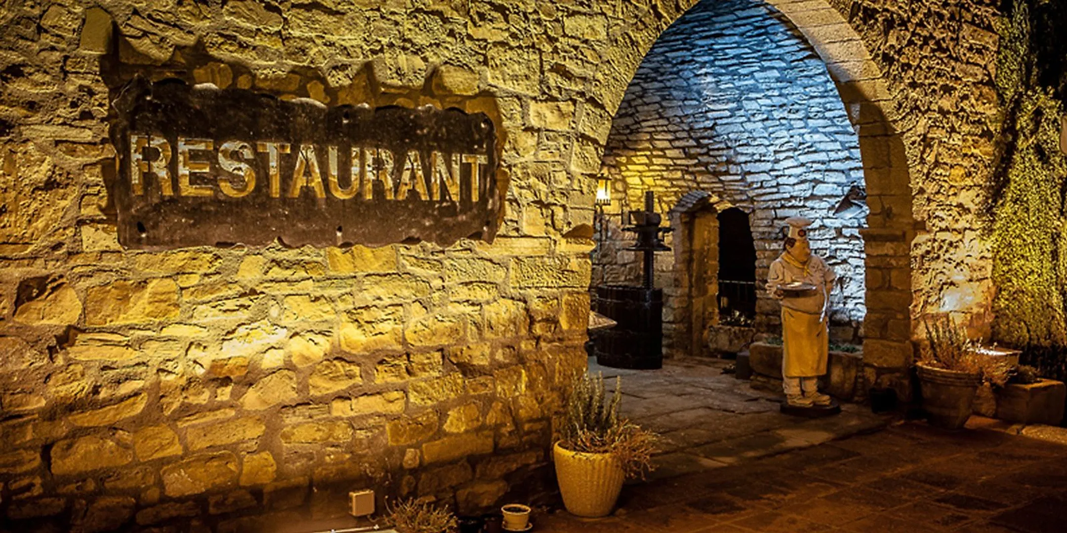 Entrada de piedra iluminada de noche, con el rótulo Restaurant en el muro y un arco que da paso al patio